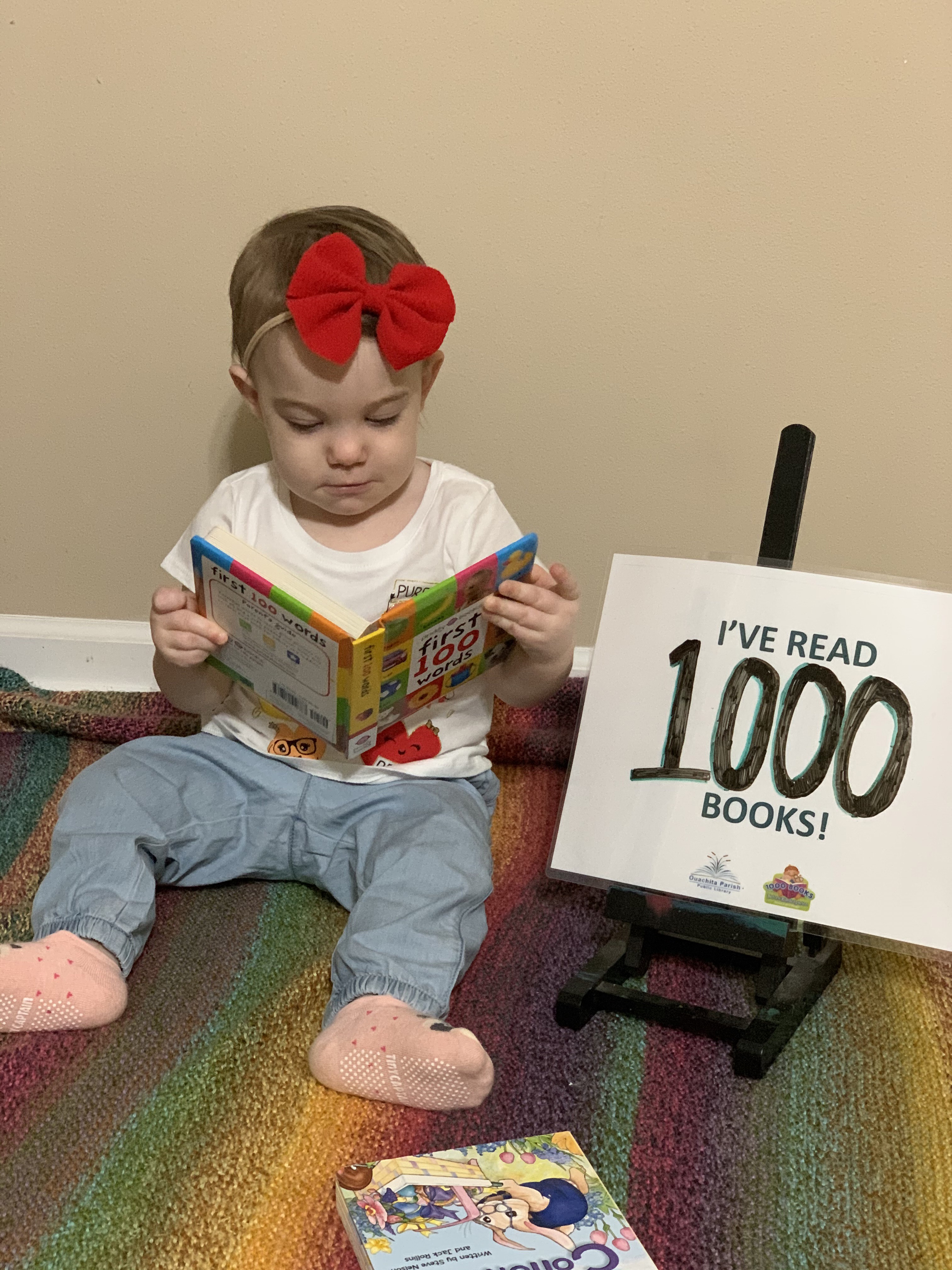 A baby sits up with a book open in her lap. There is a sign next to her that says, "I've read 1000 books!"